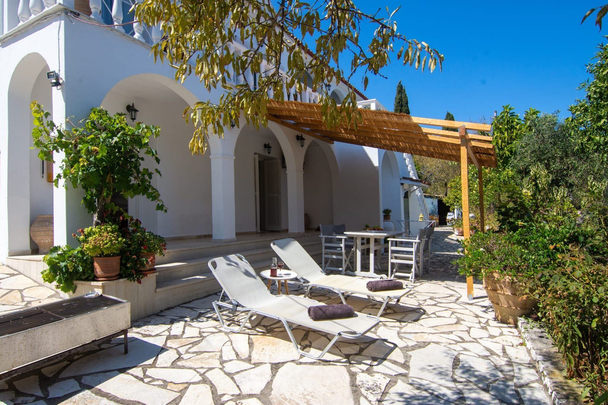 A sunlit outdoor area with stone flooring, white lounge chairs, and potted plants.
