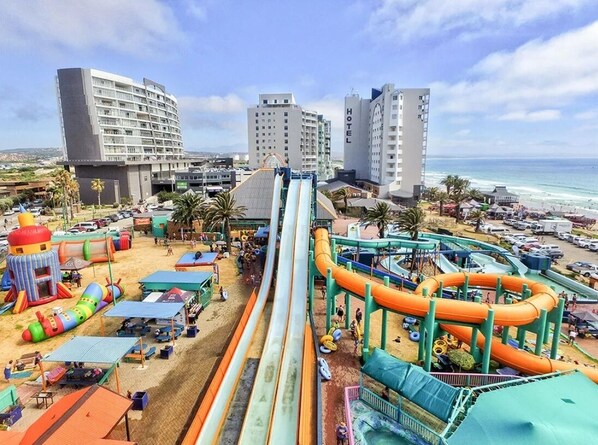 Ein Wasserpark mit mehreren Rutschen und aufblasbaren Elementen, ein Strandbereich mit Sonnenschirmen und ein Hotel im Hintergrund.