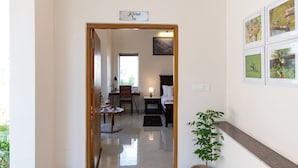 Un couloir mène à une chambre à coucher qui contient un lit, une table de chevet, une lampe et un petit bureau avec une chaise. Au-dessus de la porte, on peut voir une pancarte, et il y a une plante en pot et des tableaux encadrés accrochés au mur.