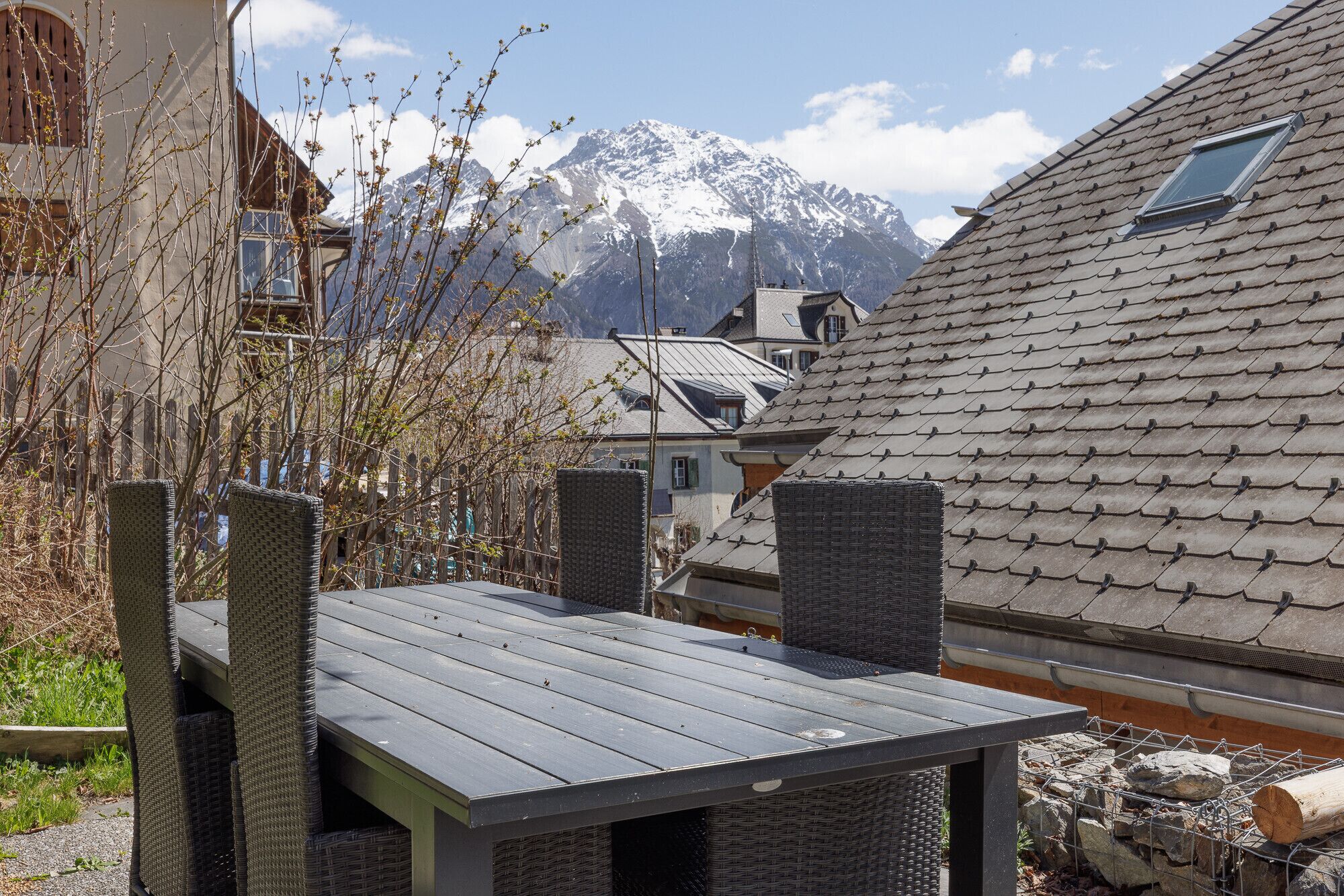 A wooden deck with wicker furniture, a mountain backdrop, and a house with a sloped roof.