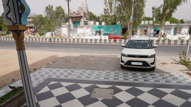 A white car parked on a checkered pavement with a manhole cover, a building with a green tarp, and a tree in the background. - Moon Mahal Hotel (Amer)