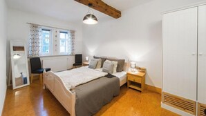 A bedroom with a wooden ceiling, a bed with a grey blanket, a bedside table with a lamp, a wardrobe, and a window with curtains.