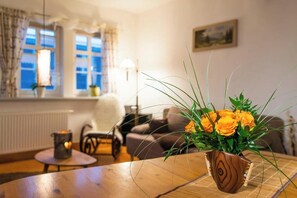 Ein Wohnzimmer mit einem Holztisch, einer Vase mit gelben Blumen, einer Lampe und einem Fenster mit Vorhängen.