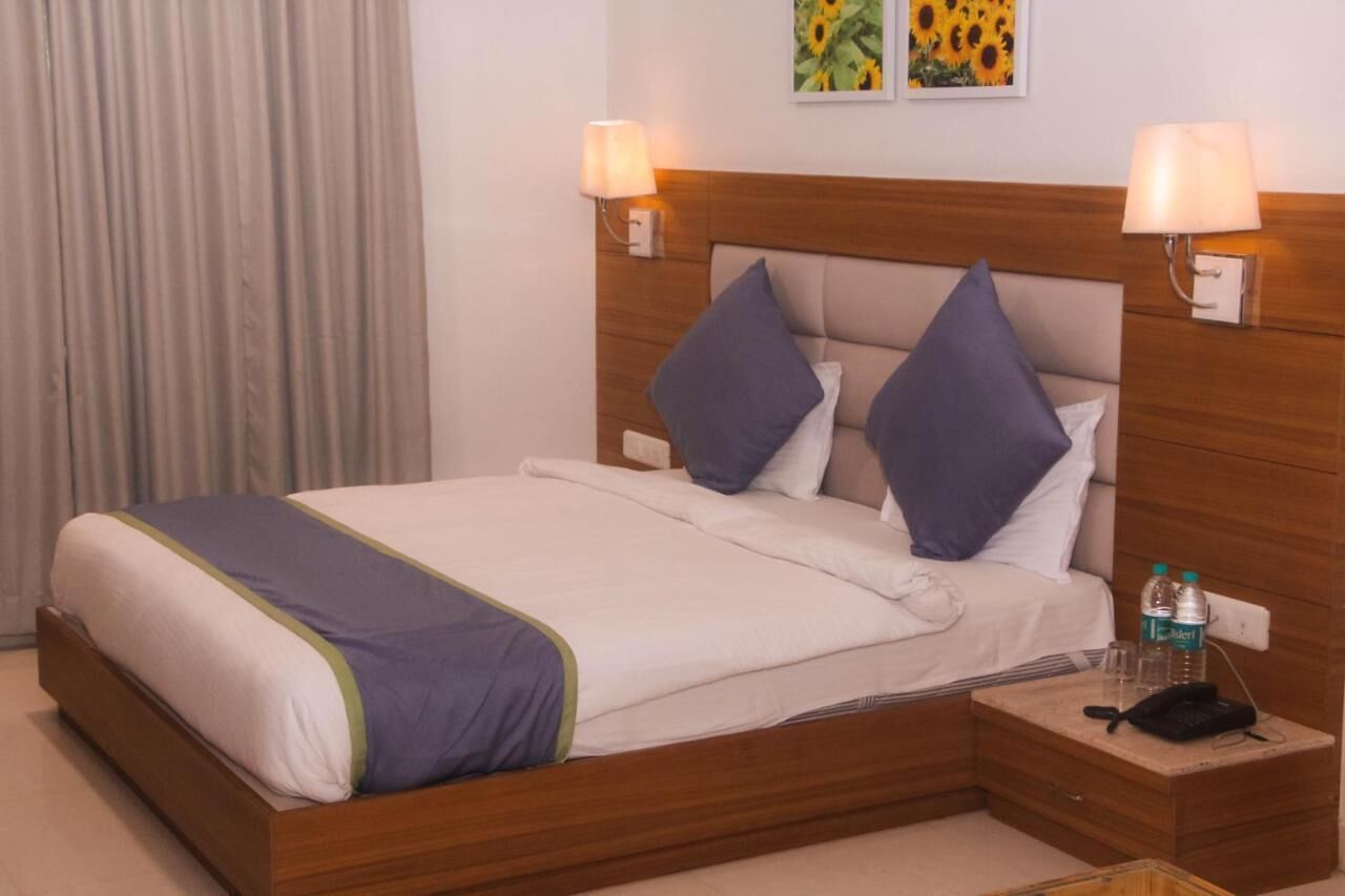A hotel room with a bed, two wall-mounted lamps, a nightstand with a phone and water bottles, and a painting above the bed.