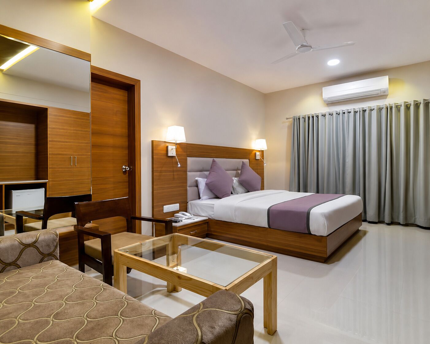 A hotel room with a bed, bedside lamps, a desk, a chair, a glass-top table, a ceiling fan, and an air conditioning unit.