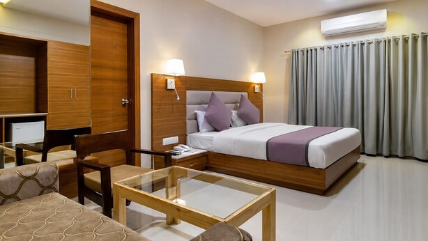 A hotel room with a bed, bedside lamps, a desk, a chair, a glass-top table, a ceiling fan, and an air conditioning unit. - HOTEL BLUE ORCHID GROUP OF MR HOTELS (Udaipur)