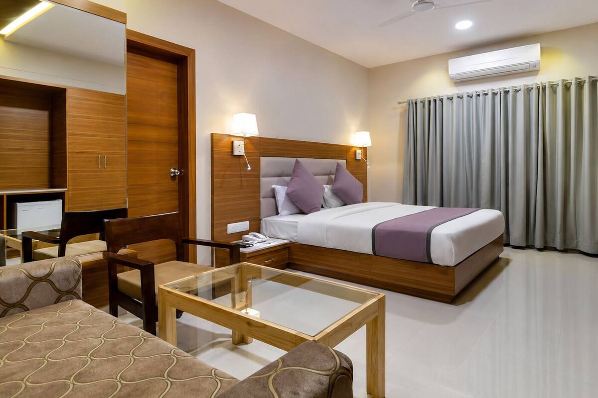 A hotel room with a bed, bedside lamps, a desk, a chair, a glass-top table, a ceiling fan, and an air conditioning unit.