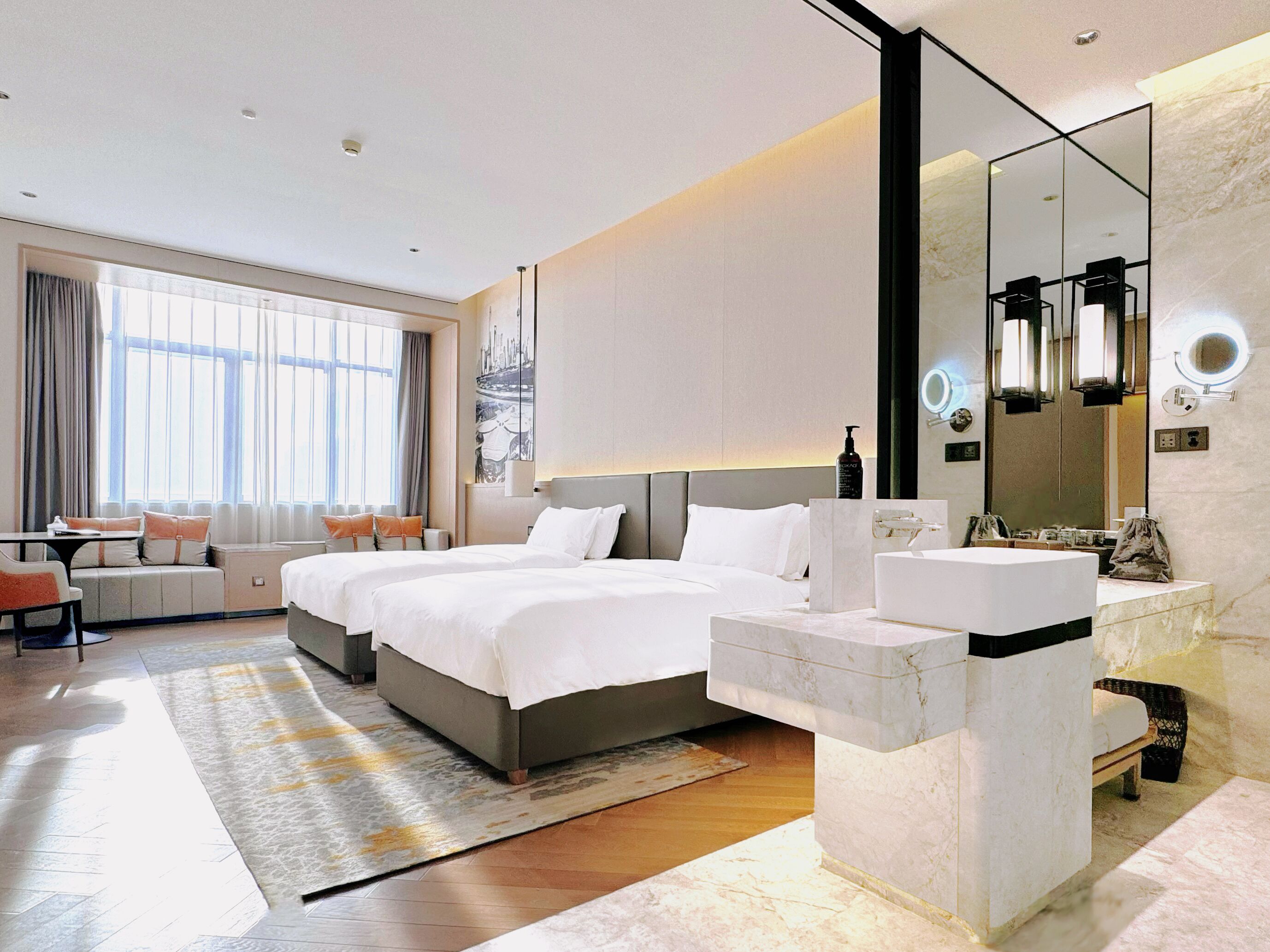 Una habitación de hotel moderna con una cama grande, un área de descanso con una mesa y sillas, un baño con un espejo amplio y lavamanos, y vistas de la ciudad a través del ventanal.