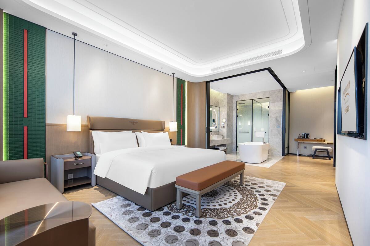 Una habitación de hotel moderna con una cama grande, mesitas de noche, un banco, un escritorio y un baño visible a través de una puerta de cristal.