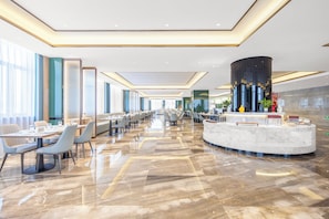 Un amplio vestíbulo de hotel con suelo de mármol, una recepción y una larga zona de comedor con mesas y sillas.