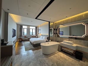 Una habitación de hotel moderna con una cama grande, una zona de estar, una bañera y un espejo amplio.