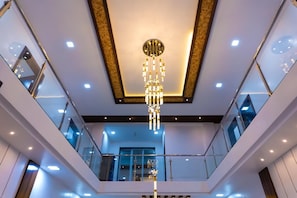 A modern interior with a central chandelier, glass railings, and a high ceiling. - Advik Farms and Resort (Ambernath)