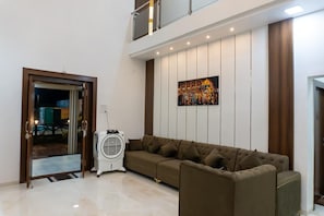 A modern living room with a sectional sofa, a standing fan, and a large wall-mounted painting. - Advik Farms and Resort (Ambernath)
