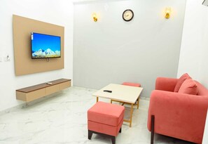 Una sala de estar moderna con una televisión de pantalla plana, un banco de madera, una mesita con un control remoto, un sofá rosa y un reloj de pared.
