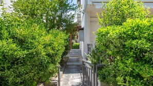 Un sendero estrecho bordeado por arbustos verdes exuberantes que conduce a un edificio de fachada blanca y un balcón.