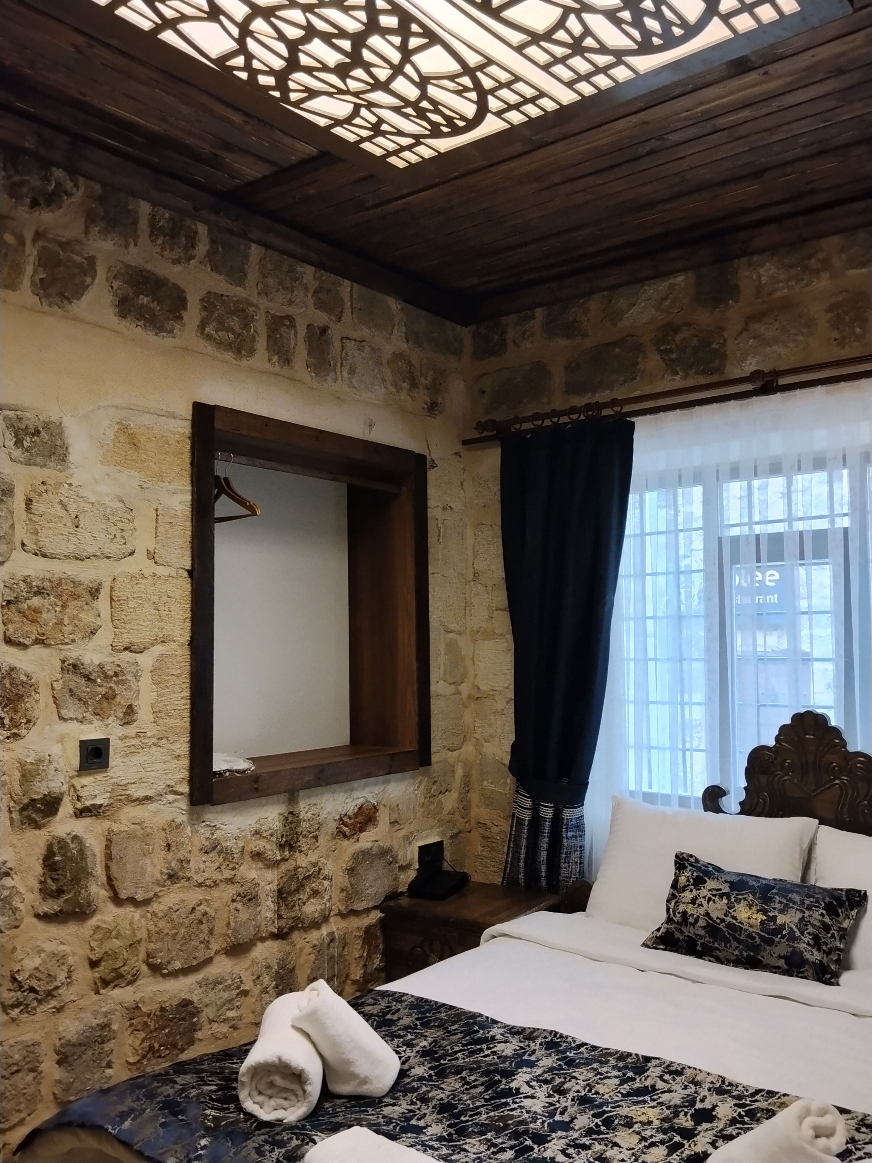 Una habitación con paredes de piedra, techo de madera, una cama con colcha estampada, un perchero y una ventana con cortinas.