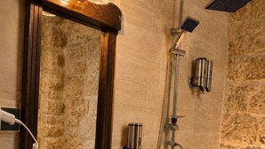 Casa de banho com parede de pedra, pia redonda, espelho com luzes, chuveiro com chuveiro manual e uma pequena planta em vaso.