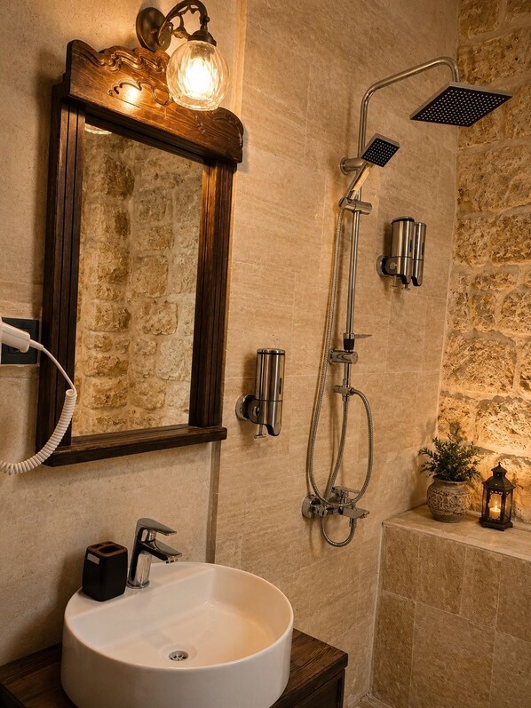 A bathroom with a stone wall, a round sink, a mirror with lights, a shower with a handheld showerhead, and a small potted plant. - Kasrı Hatun Konağı (Mardin)