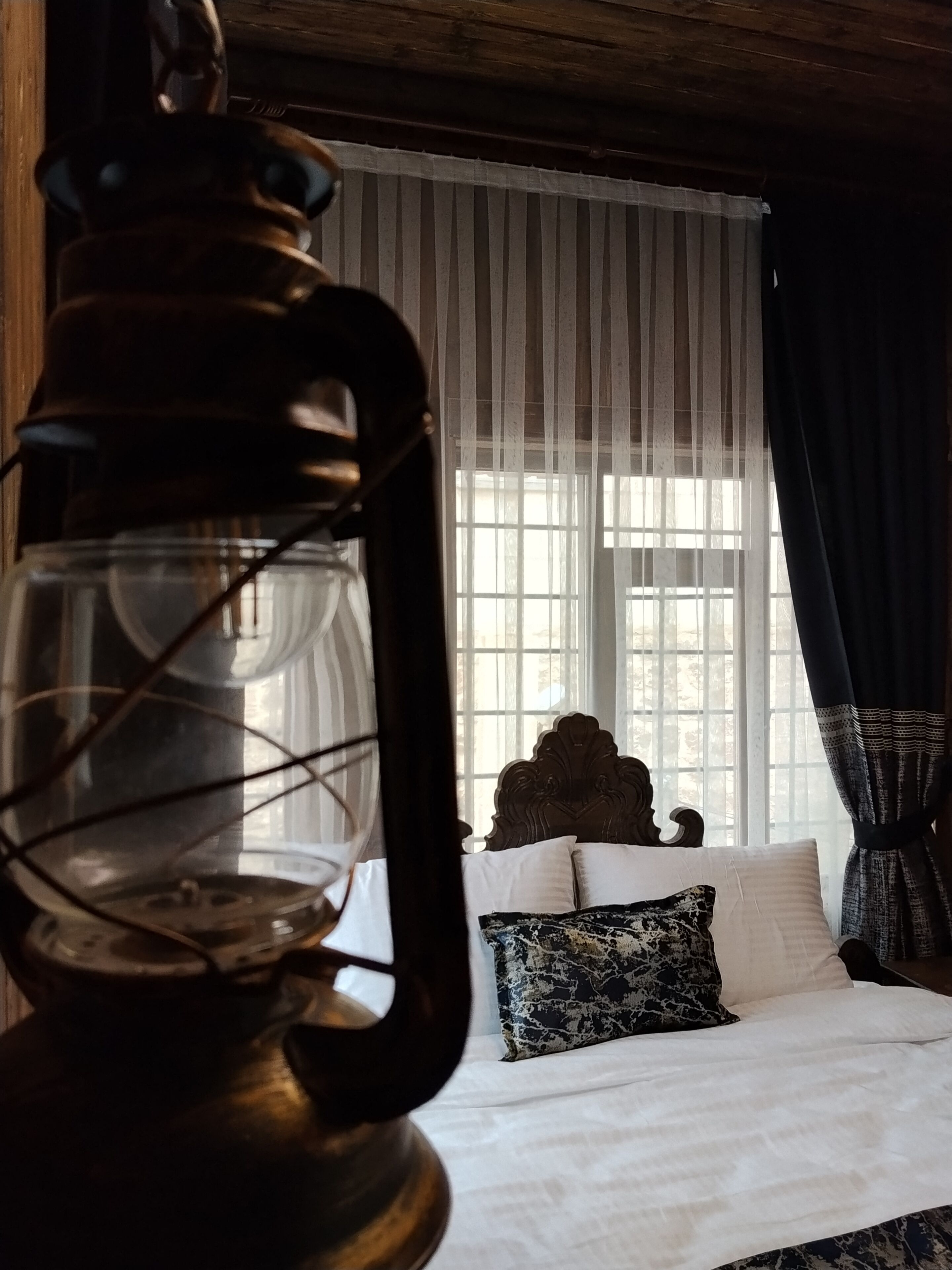 Une pièce avec un lit, une fenêtre ornée de rideaux légers et une lampe de couleur sombre placée au premier plan.
