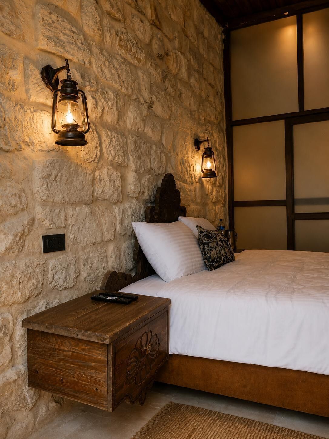 Un dormitorio con pared de piedra, mesita de noche de madera, cama con ropa de cama blanca y dos faroles fijados a la pared.