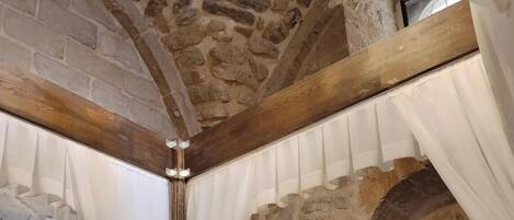 A stone-walled room with a wooden ceiling, a bed with white bedding, and a chandelier.