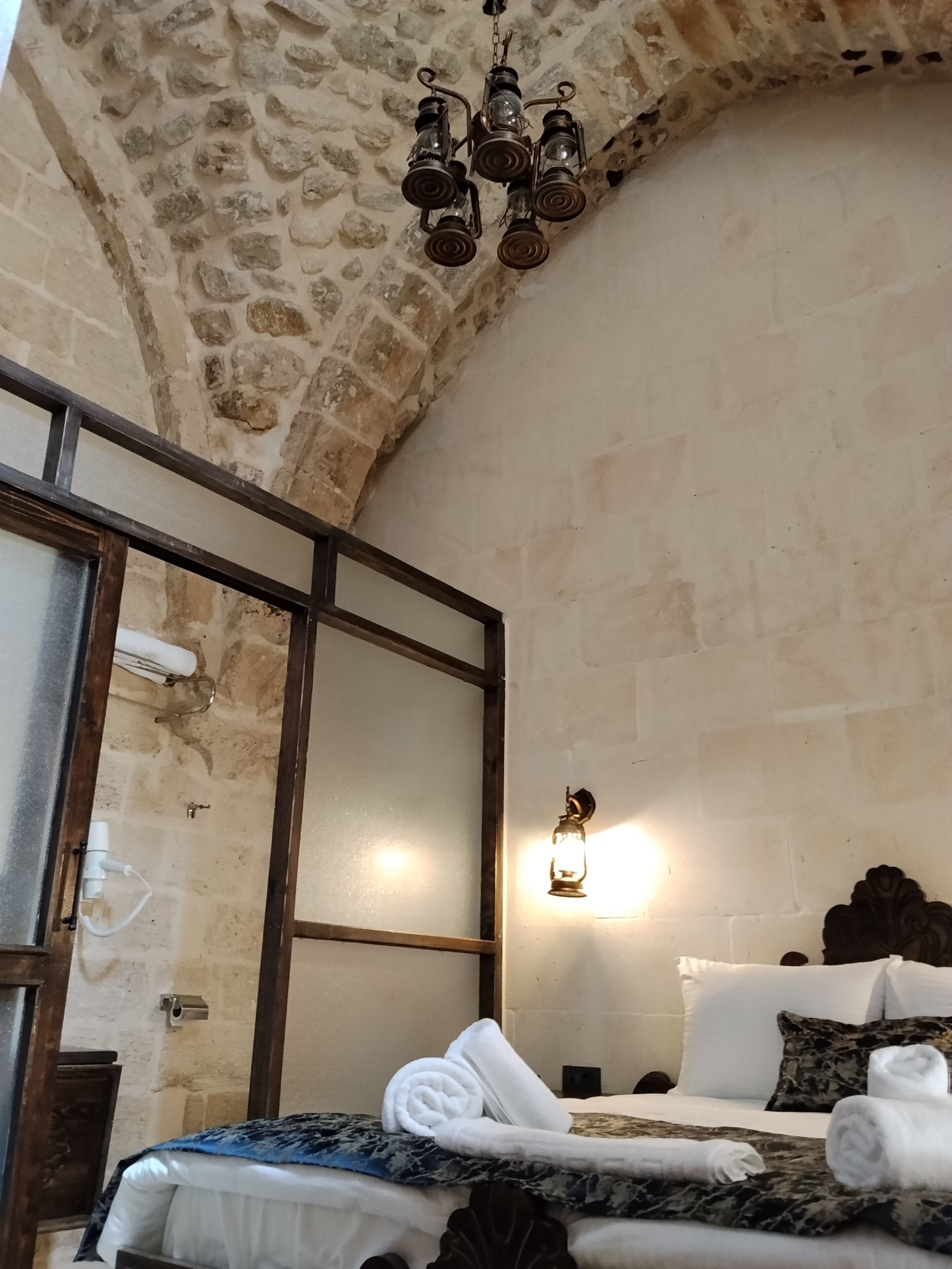 Une chambre d’hôtel avec des murs en pierre, un lit avec une literie blanche et des serviettes roulées, une tête de lit en bois, un luminaire suspendu et un grand miroir.