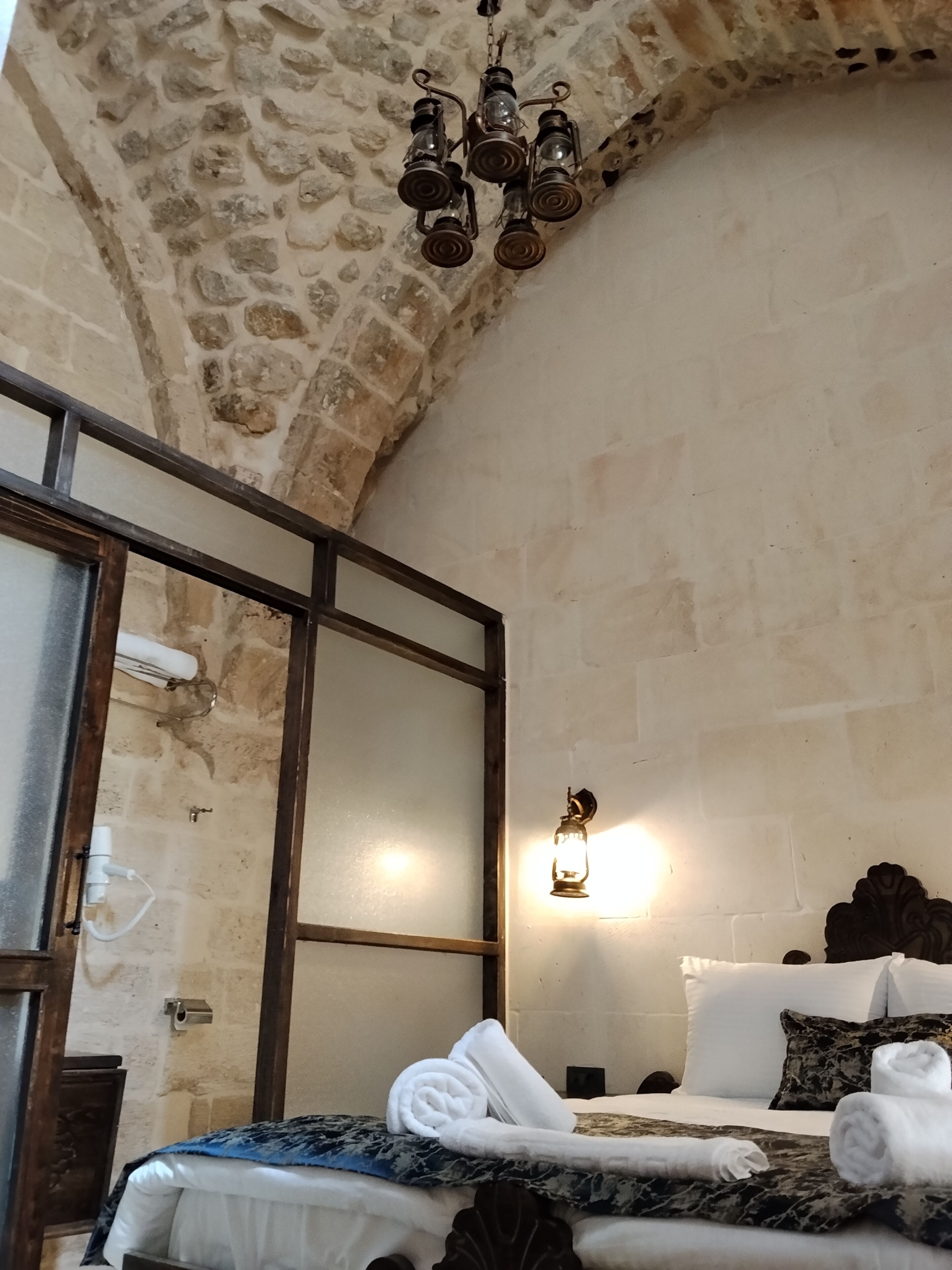A room with a stone wall, a bed with white bedding and a blue patterned cover, a wooden headboard, a hanging light fixture, and a wall-mounted light.