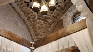 Quarto com paredes de pedra, uma cama, cortinas brancas e teto de madeira.