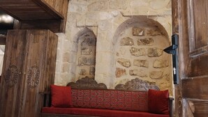 Quarto com parede de pedra, vigas de madeira, um banco com almofadas vermelhas e um lustre pendurado.