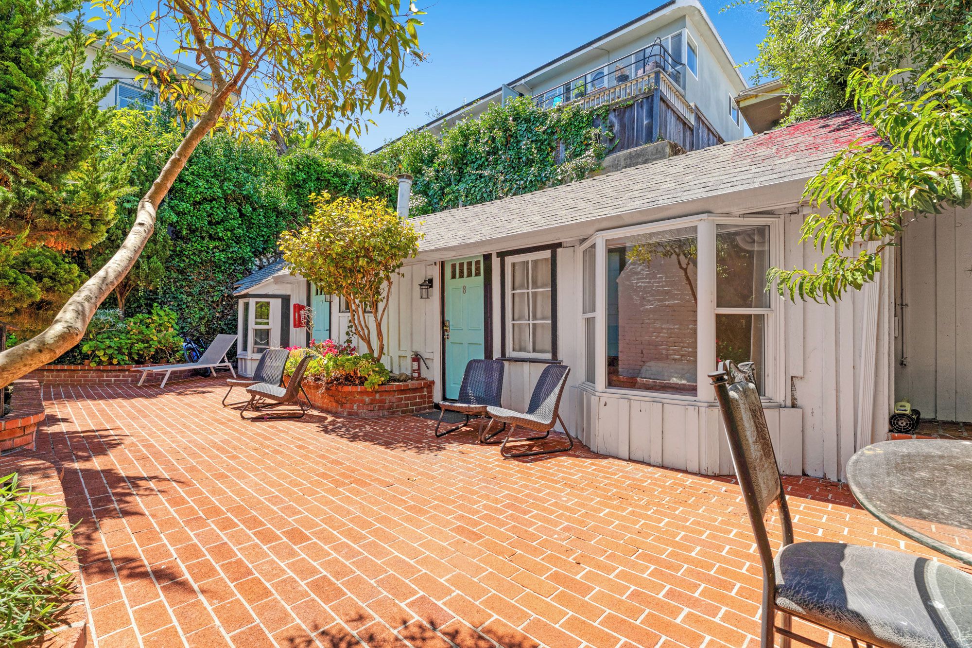 A patio with brick flooring, a white building with a window, and two chairs.