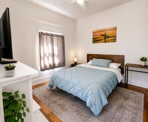 A bedroom with a bed, a TV on a stand, a window with curtains, a painting on the wall, and a bedside table with a lamp. - charming 1 bed-private bath great location by Beach, Pier, Boardwalk-fast WiFi-8 (Santa Cruz)