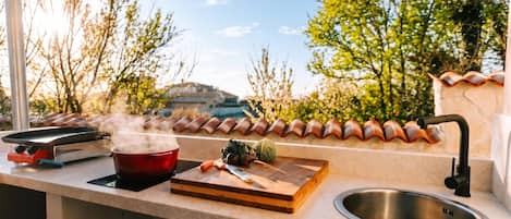 An outdoor kitchen with a gas stove, a red pot, a cutting board with vegetables, and a sink.