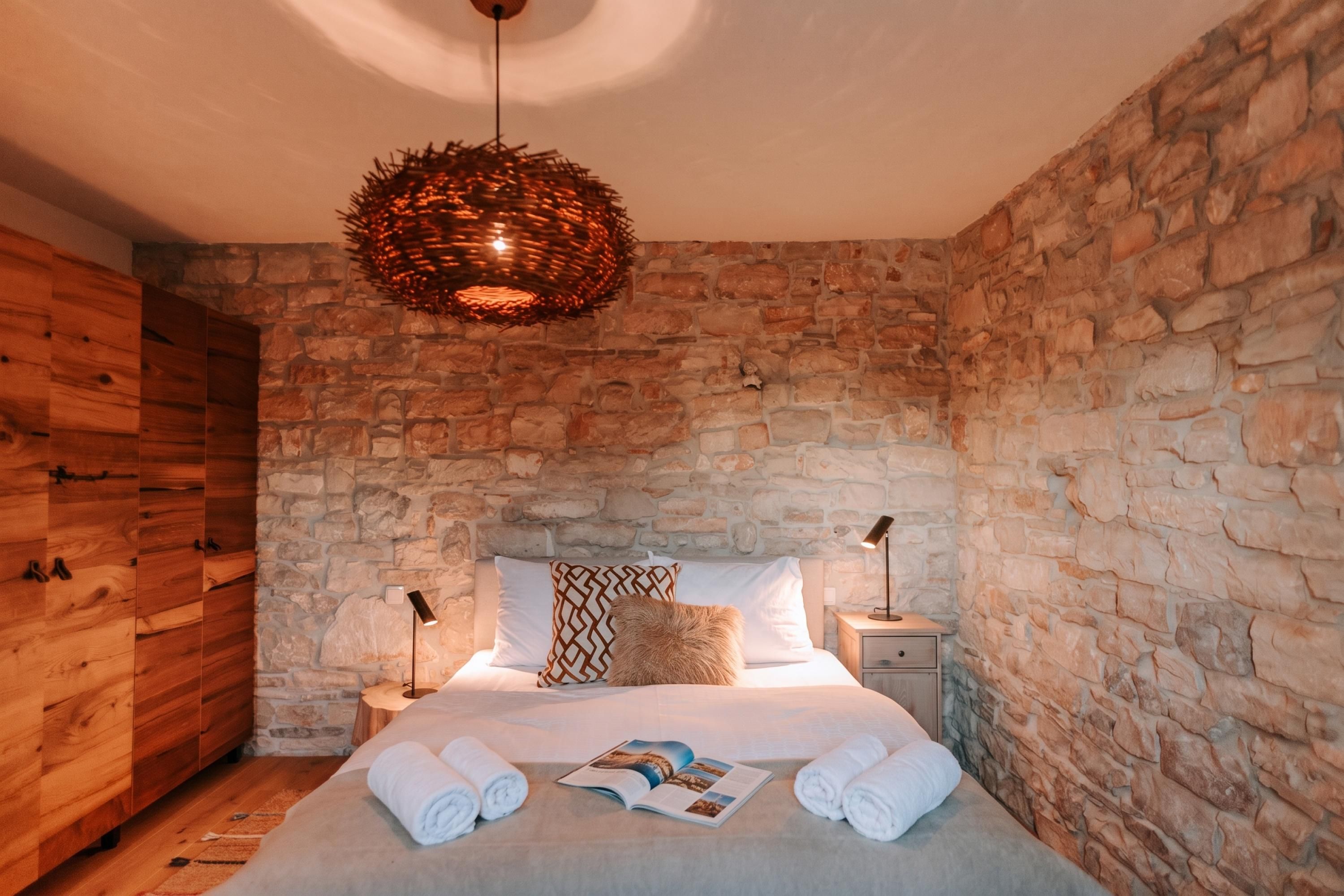 Un dormitorio con pared de piedra, un armario de madera, una cama con ropa blanca, una lámpara decorativa colgante y una revista sobre la cama.