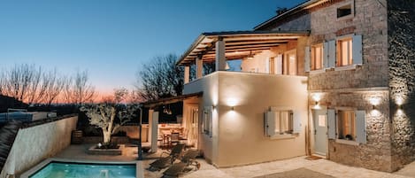 Una casa de piedra de dos plantas con piscina, mobiliario de exterior y un porche cubierto.