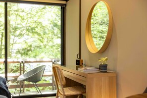 A room with a wooden desk, a chair, a round mirror, a potted plant, and a view of greenery outside.