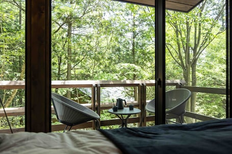 A room with a bed, two chairs, a small table, and a view of a forest through large windows.. Iida Highlands is located near Achi Village, the home of Japan's starry sky. A room in a hideaway where the scent of the forest and the star-filled sky will soothe you.