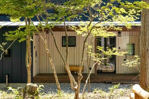 Un bâtiment moderne aux murs extérieurs en bois, doté d’une terrasse en bois et surmonté d’un grand arbre, visible au premier plan.