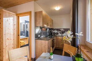 A compact kitchen with wooden cabinets, a stainless steel range hood, and a dining area with a table and chairs. - Refugium Dolomiten & Chalet (Sillian)