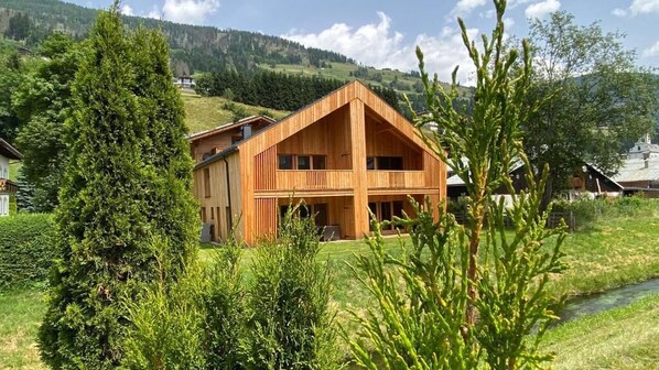 A two-story wooden house with a balcony, surrounded by greenery and a river. - Refugium Dolomiten & Chalet (Sillian)