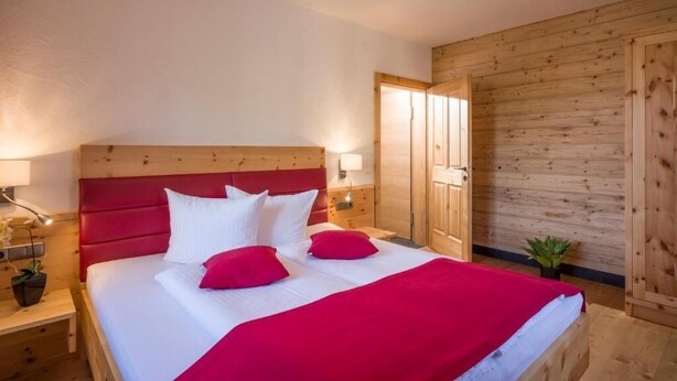 A bedroom with a wooden headboard, a bed with white and red bedding, a nightstand with a lamp, and a wooden wardrobe. - Refugium Dolomiten & Chalet (Sillian)