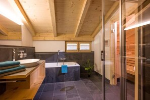 A modern bathroom with a wooden ceiling, a freestanding bathtub, a glass shower enclosure, and a wooden vanity with a sink and towels. - Refugium Dolomiten & Chalet (Sillian)