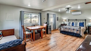 Una habitación con piso de madera, una cama con estructura metálica, una mesa de comedor con sillas, un refrigerador y una ventana con cortinas.