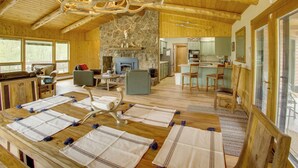 Un interior espacioso de madera con chimenea de piedra, comedor con mesa dispuesta para cuatro, y cocina visible a través de un vano abierto.
