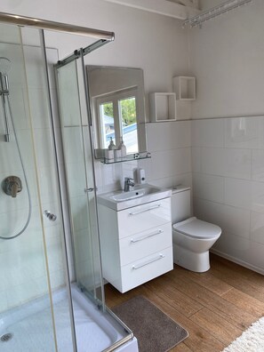 Ein modernes Badezimmer mit einer Duschkabine aus Glas, einem weißen Waschbecken mit Spiegel darüber, einer Toilette und einem kleinen Ablagebereich für Toilettenartikel.