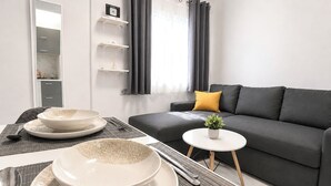 A modern living room with a grey sofa, a small white table, and a dining area with plates and bowls. - Athens Explorer Studio Near Transit by ΜΡS (Agios Ioannis Rentis)