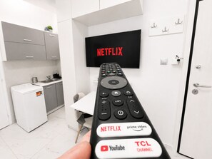 A modern kitchen with a washing machine, a TV mounted on the wall displaying 'NETFLIX', and a remote control with buttons for Netflix, Prime Video, YouTube, and TCL CHANNEL.