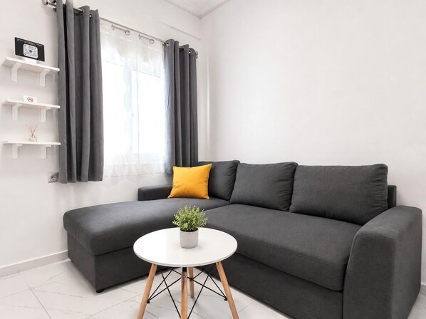 Un salon moderne avec un canapé gris, une petite table blanche ornée d’une plante, des rideaux gris et des étagères blanches.