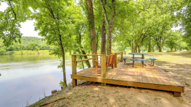 Eine Holzterrasse mit Bänken und einem Picknicktisch am Ufer eines ruhigen Sees, umgeben von Bäumen.