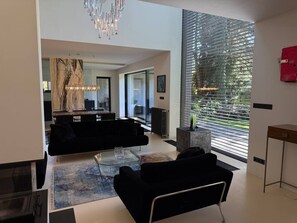 A modern living room with a black sofa, a glass coffee table, and a chandelier. There is a large window with blinds and a view of greenery outside.