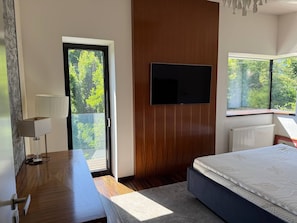 A modern bedroom with a bed, a TV mounted on the wall, a desk with a lamp, and a view of greenery through the window.